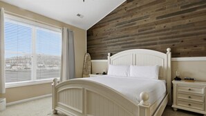 Ein Schlafzimmer mit großem Fenster, einer Holzwand als Blickfang, einem weißen Bett und einem Nachttisch mit Schubladen.
