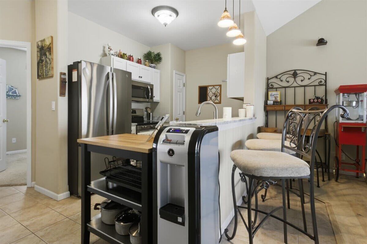 A modern kitchen with a stainless steel refrigerator, a water dispenser, and a coffee machine. There are two bar stools and a small island with a sink.