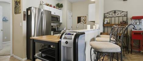 A modern kitchen with a stainless steel refrigerator, a water dispenser, and a coffee machine. There are two bar stools and a small island with a sink.