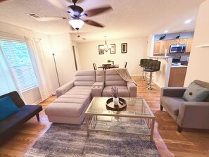 Ein Wohnzimmer mit einer Couch, einem Glastisch, einem Deckenventilator und einer Küchenzeile mit Barhocker.