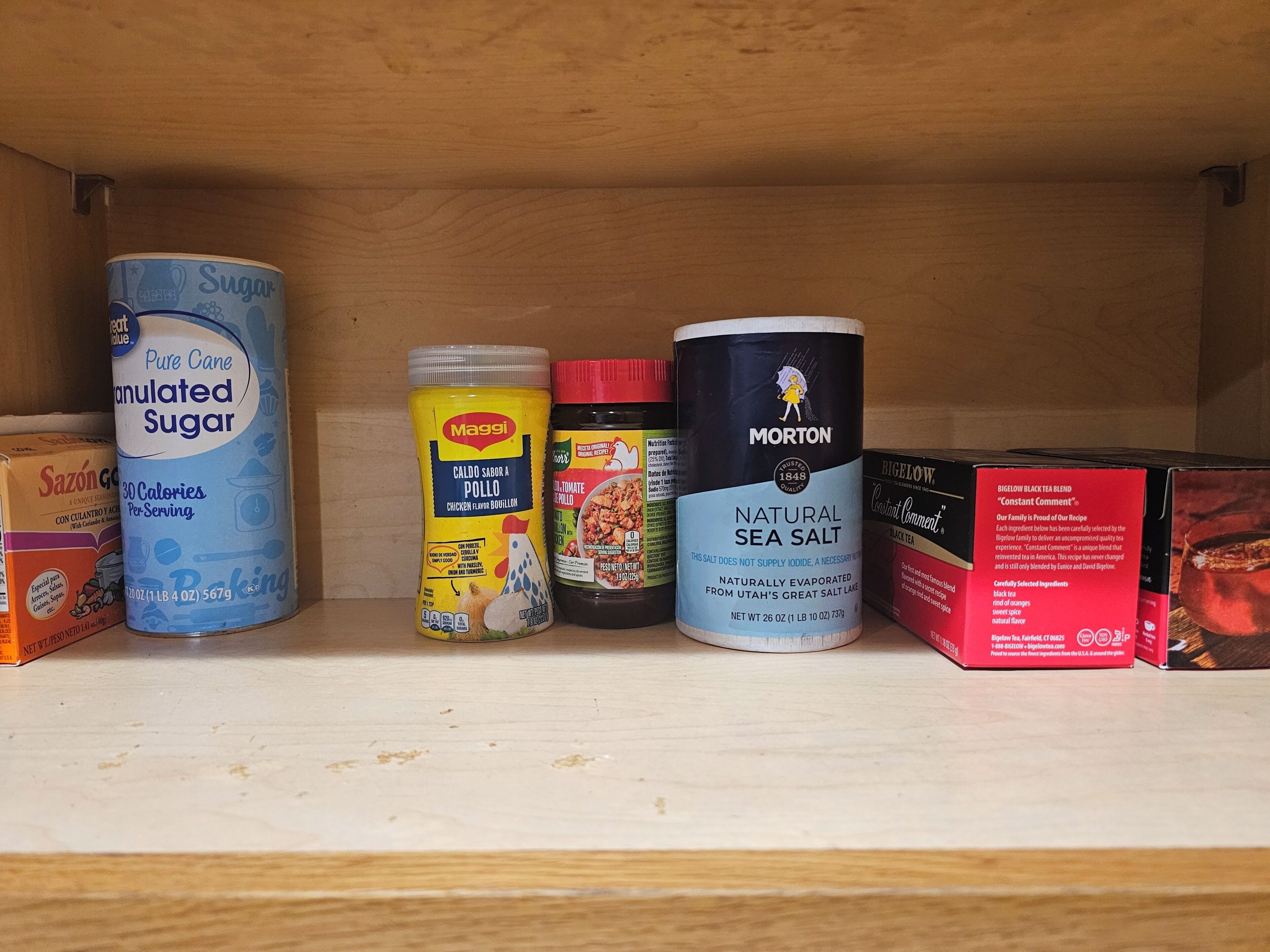 A shelf with various food items including a can of sugar, a jar of chicken flavoring, a container of sea salt, and a box of tea.