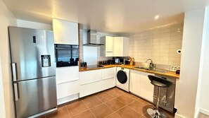 A modern kitchen with a stainless steel refrigerator, white cabinets, a washing machine, and a bar stool. - Cosy London Apartment Near City Centre (London)