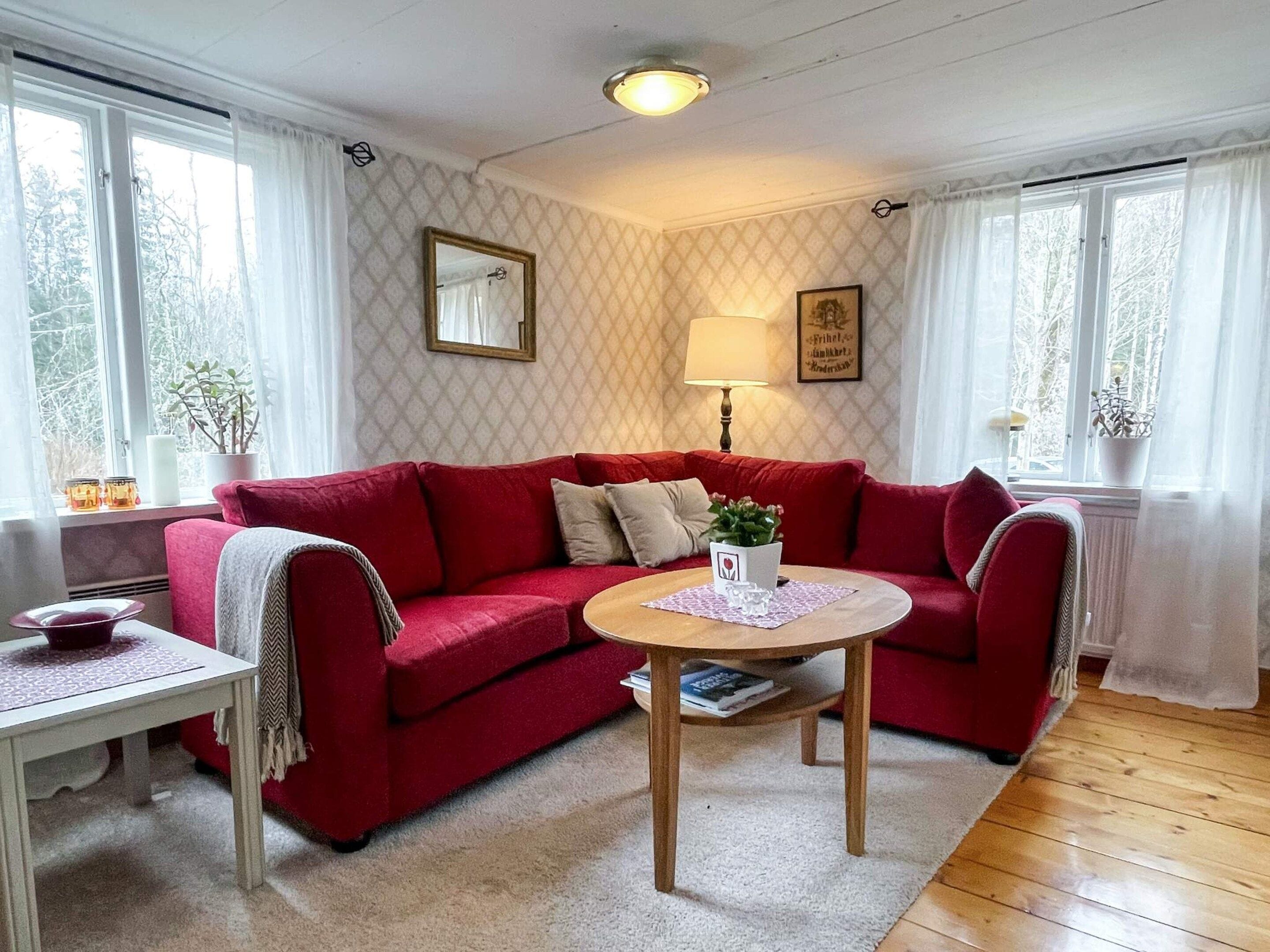 A cozy living room with a red sofa, a small round table, and a lamp. There are windows with curtains, a mirror on the wall, and a potted plant.