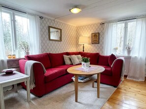 Ein gemütliches Wohnzimmer mit einem roten Sofa, einem kleinen runden Tisch und einer Lampe. Es gibt Fenster mit Vorhängen, einen Spiegel an der Wand und einen Blumentopf.