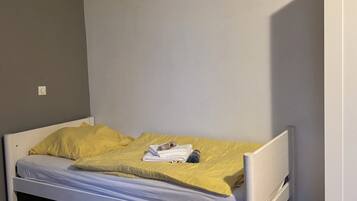 Un lit simple avec une couverture jaune et des serviettes pliées, un plancher en bois et une armoire blanche.