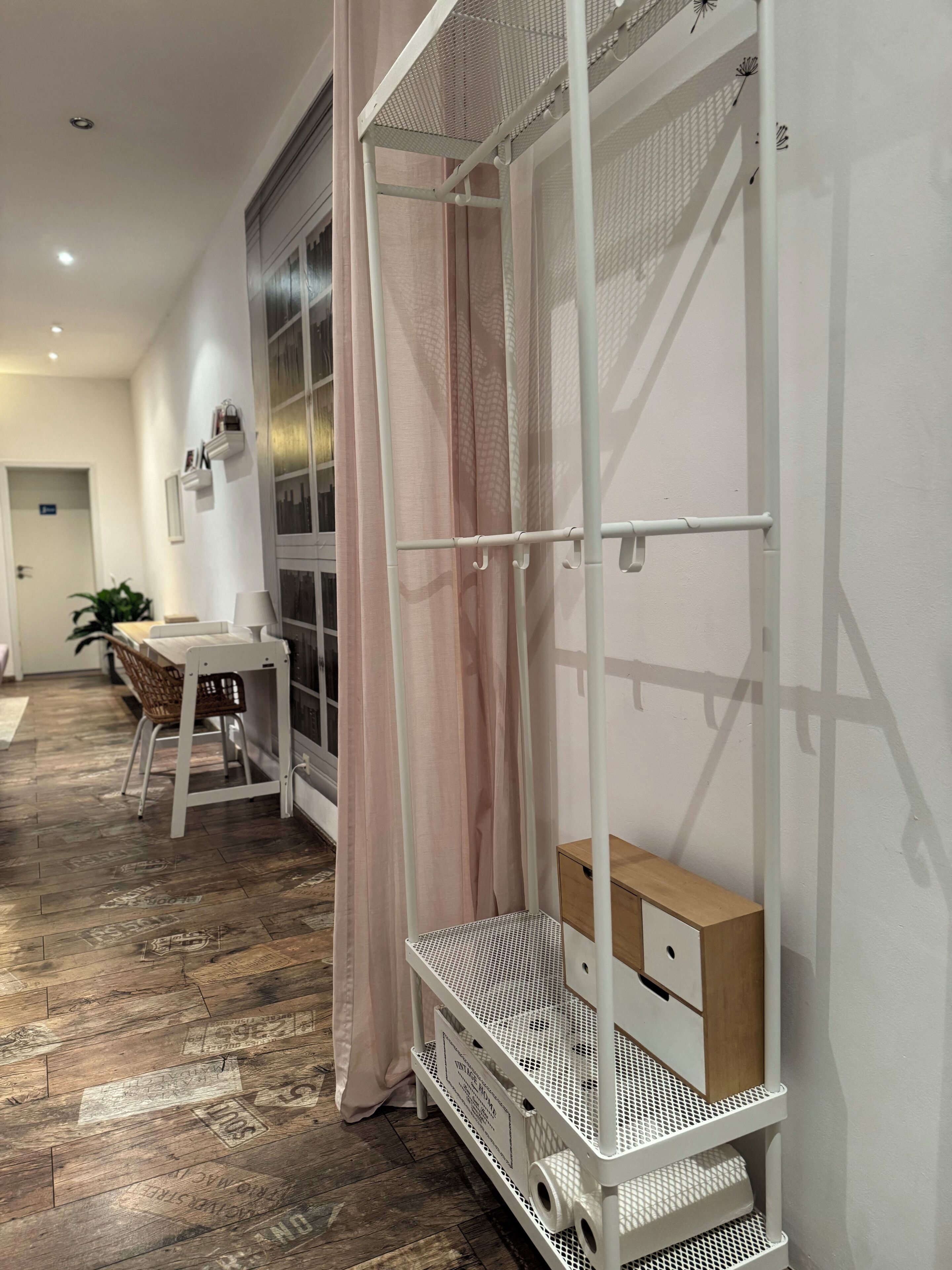 Ein Zimmer mit einer weißen Metallregalkonstruktion, einem Holztisch, einem Korbstuhl und einem Fenster mit transparenten Vorhängen.