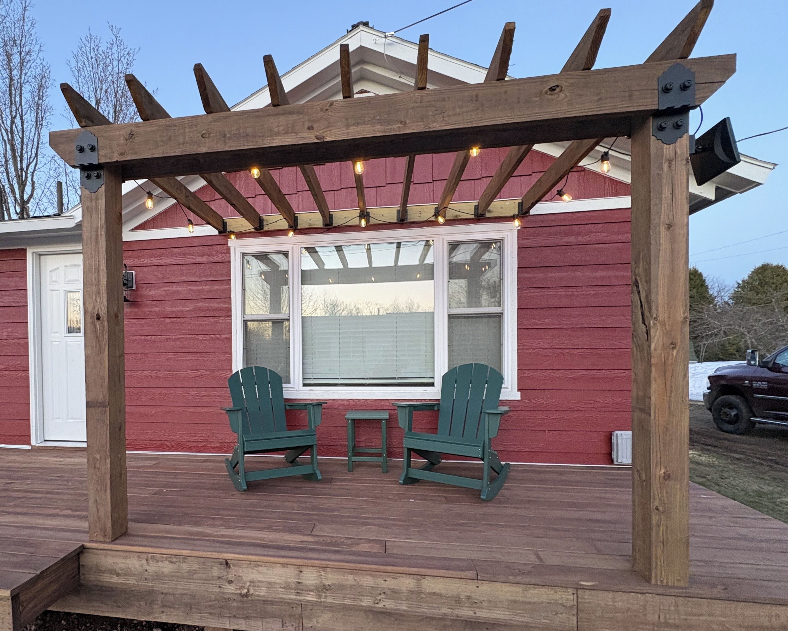 A wooden pergola with string lights, two green Adirondack chairs, and a table on a wooden deck.