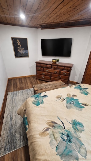 A bedroom with a wooden ceiling, a bed with floral bedding, a dresser, a flat-screen TV, and a framed picture on the wall.