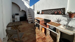 Un patio con una mesa de madera, un banco, un horno de ladrillo y una pequeña zona de cocina con un fregadero y plantas en macetas.