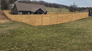 A house with a wooden fence, a grassy yard, and a hilly landscape in the background.