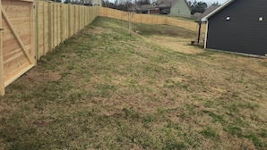 A residential backyard with a wooden fence, a grassy slope, and houses in the background.