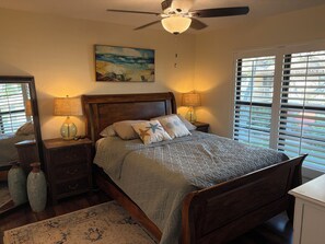 A bedroom with a wooden bed, two bedside tables with lamps, a ceiling fan, a beach-themed painting, and a view through large windows. - Great beach access, central location, 2-bedroom condo in wonderful Marco Island (Marco Island)