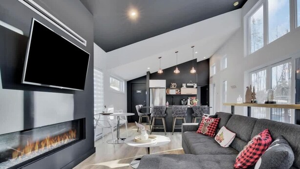 A modern living room with a fireplace, a TV mounted on the wall, a grey sofa with red and black pillows, and a dining area with a bar. - Atlas | Familial | Piscine & Spa by Alchalet (Pont-Rouge)