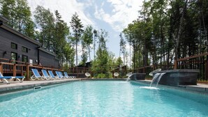 A swimming pool with lounge chairs and a waterfall feature, surrounded by trees and a building. - Atlas | Familial | Piscine & Spa by Alchalet (Pont-Rouge)