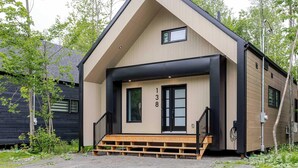 Una casa moderna de dos plantas con un techo a dos aguas destacado, un porche con escalera de madera y una puerta con marco negro.