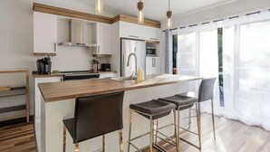 Una cocina moderna con una isla central, taburetes de bar, iluminación colgante y ventanas amplias.