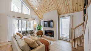 A modern living room with a high ceiling, a fireplace, a TV, and a staircase. - ØM de Portneuf - Spa privé - Nature by Alchalet (Pont-Rouge)