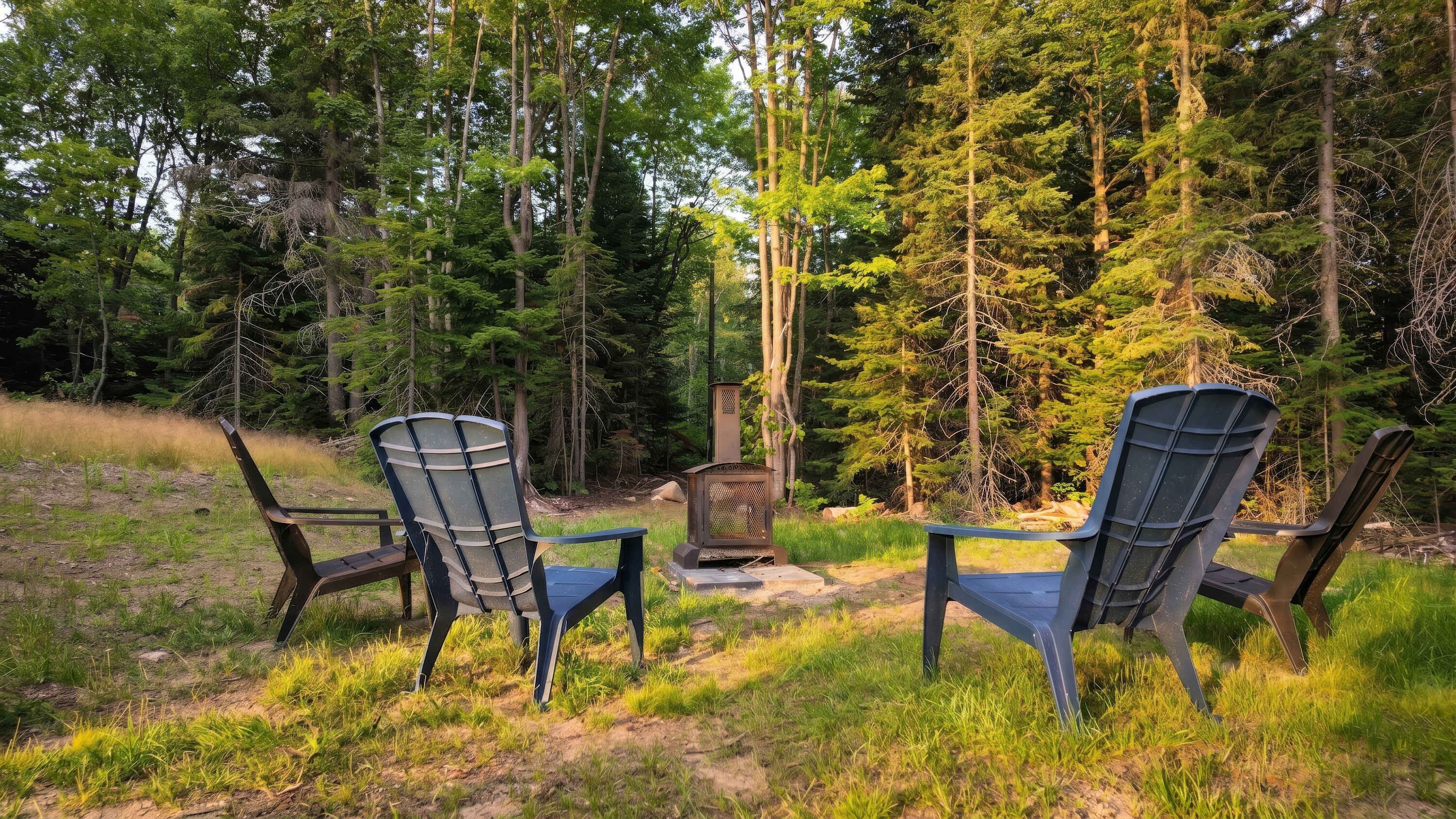 Two Adirondack chairs facing a wood-burning stove in a forested area.