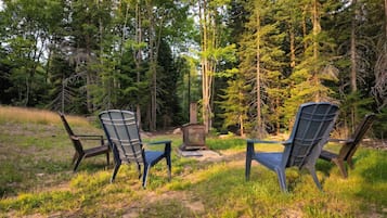 Two Adirondack chairs facing a wood-burning stove in a forested area.