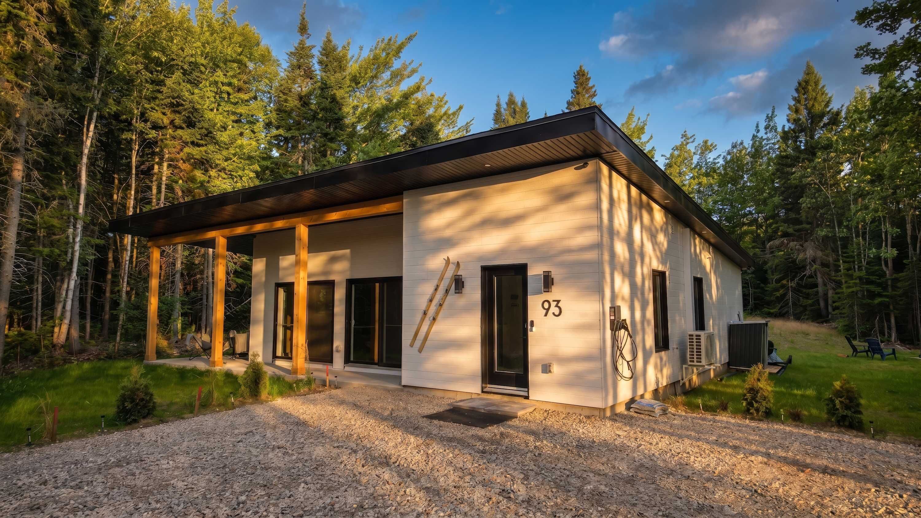 A modern cabin with a covered porch, a ski rack, and a gravel driveway.