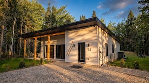 A modern cabin with a covered porch, a ski rack, and a gravel driveway.