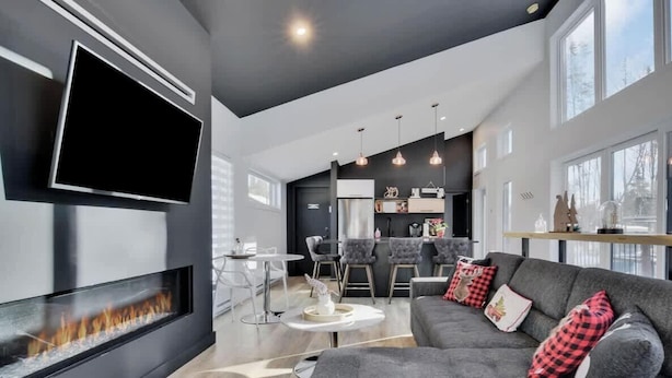 A modern living room with a fireplace, a TV mounted on the wall, a grey sofa with red and black pillows, and a dining area with a bar.