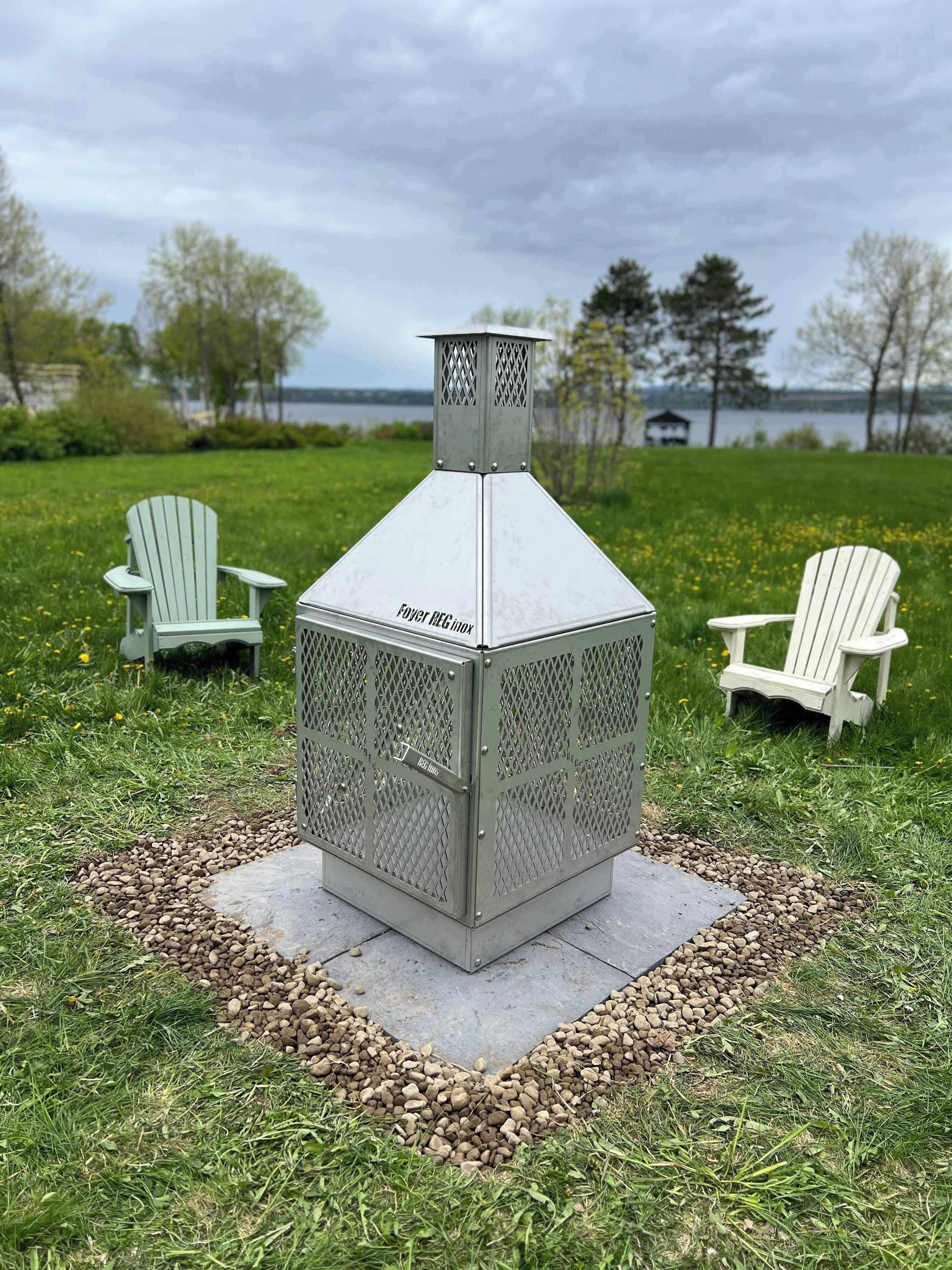 A metal outdoor grill with a chimney, surrounded by a stone border, with two Adirondack chairs and a lake view in the background.