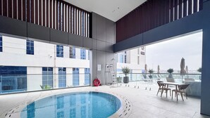 A modern indoor swimming pool with a view of the cityscape, a lounge area with tables and chairs, and a lifebuoy near the pool. - PVH - Royal Regency - Modern Studio (Dubai)