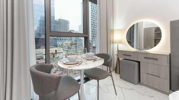 A modern hotel room with a dining table set for two, a round mirror, and a view of the cityscape through large windows. - PVH - Royal Regency - Modern Studio (Dubai)