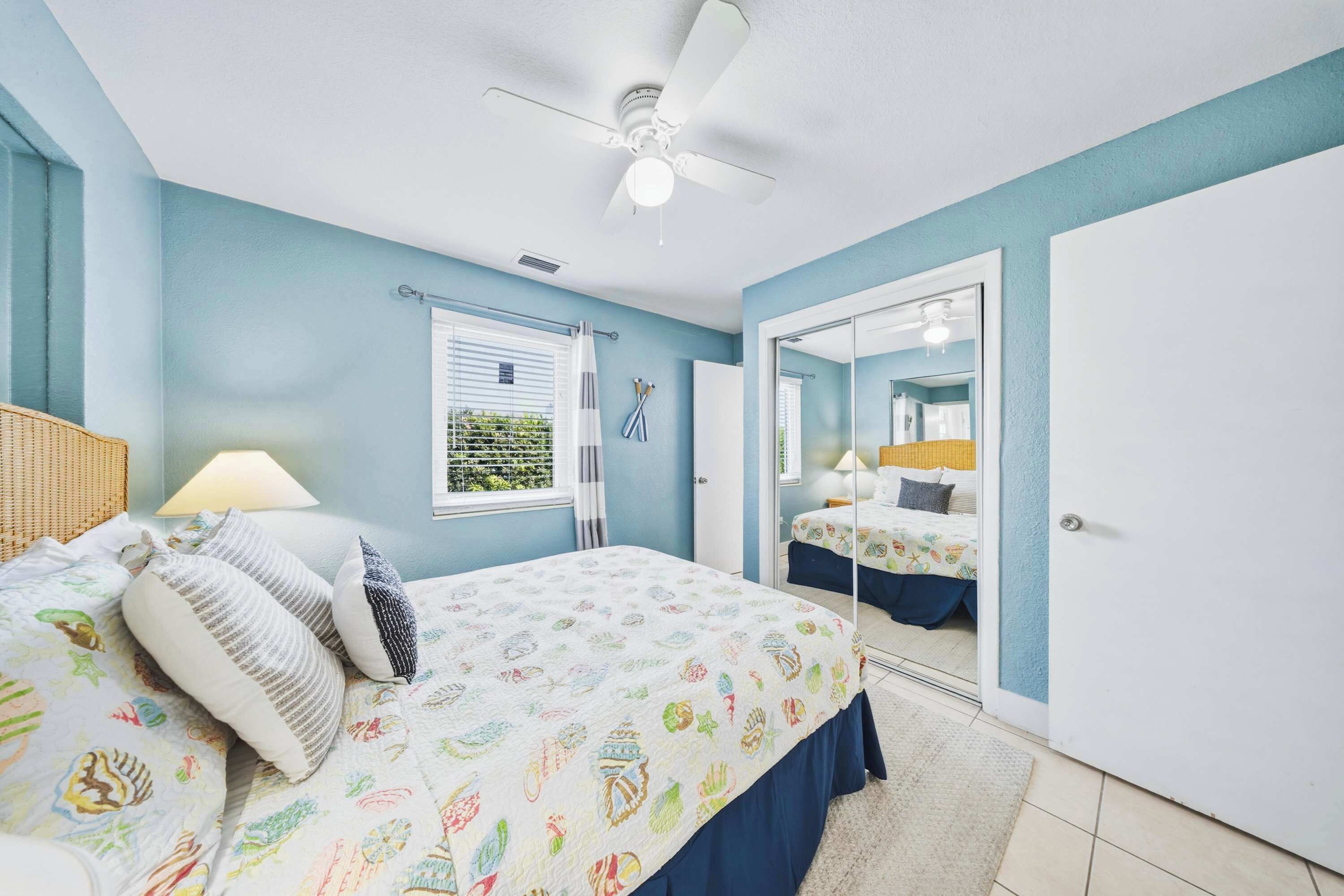 A bedroom with a bed, a ceiling fan, a window with blinds, a mirror, and a door leading to another room.