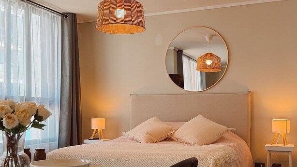 A bedroom with a bed, bedside tables, a round mirror, a dining table with chairs, and a vase of flowers. - Escapada en Valparaíso (Valparaíso)