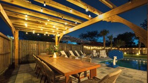 Un patio al aire libre con una mesa y sillas de madera, una piscina y luces colgantes.