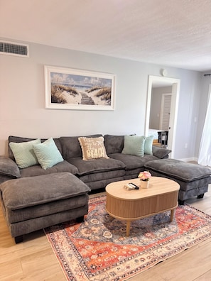 Ein Wohnzimmer mit einer Eckcouch, einem Couchtisch, einem gemusterten Teppich und einem Strandgemälde.