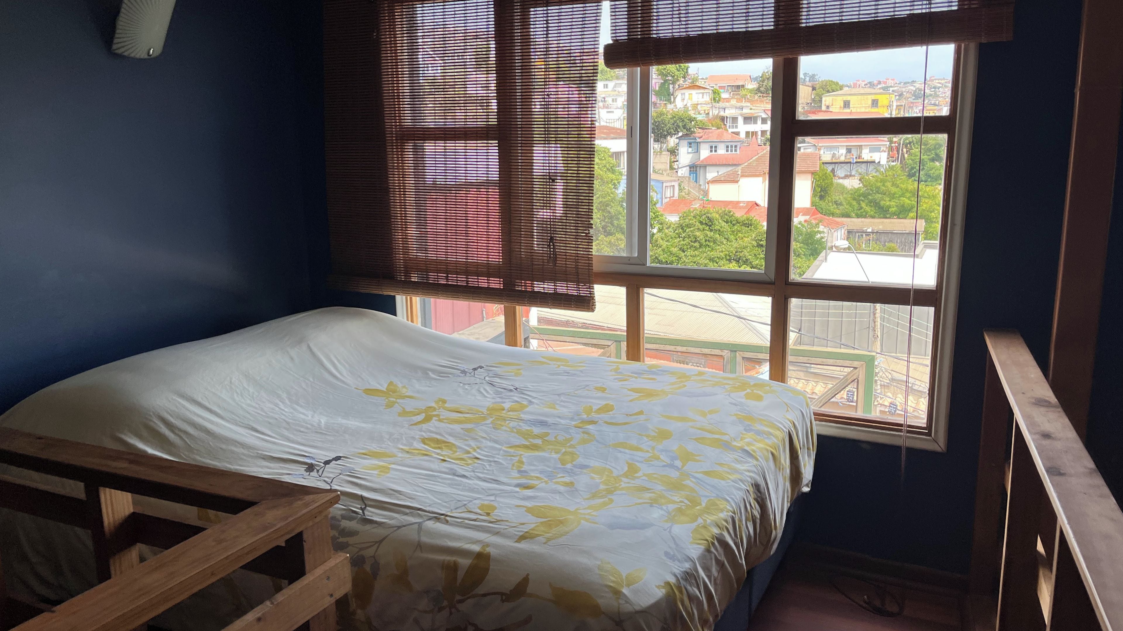 Une chambre avec un lit, une fenêtre avec des stores et une vue sur les bâtiments environnants.