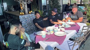 Un grupo de personas cenando en una mesa al aire libre con mantel morado, rodeado de platos y cubiertos.