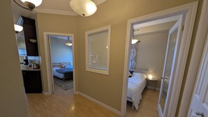 Corredor com uma cama, uma mesinha e uma porta que dá para outro quarto.