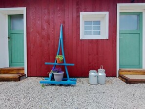 Un bâtiment en bois rouge avec deux portes vertes, une petite fenêtre et un présentoir bleu orné de plantes en pot.