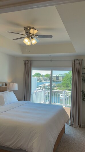 A hotel room with a bed, a ceiling fan, a nightstand, a lamp, and a view of a marina and buildings through large windows. - Waterfront • Heated Decks • Workspace • EV Charging • Home Gym • Downtown (Cambridge)