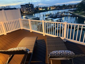 A wooden deck with two wicker chairs, a railing, and a view of a marina with boats and buildings in the background. - Brand New Waterfront Home • Pool • Walk Downtown & Events • Sleeps 8 (Cambridge)