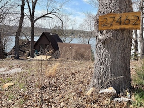 A rustic cabin with a wooden number sign, surrounded by trees and a lake.