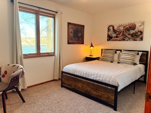 A bedroom with a bed, a nightstand, a lamp, a chair, a window with curtains, and two paintings on the wall.