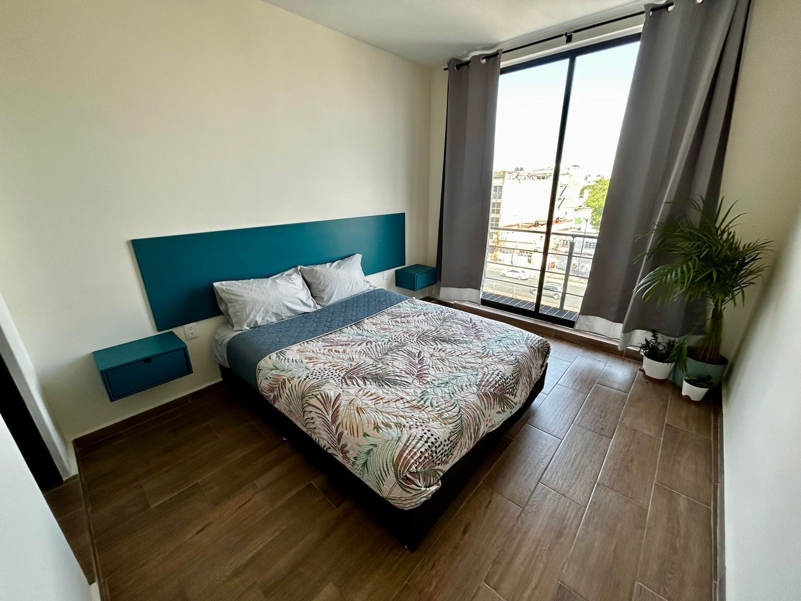 A bedroom with a bed, a nightstand, a window with curtains, and a potted plant.