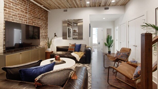 A living room with a brick wall, a flat-screen TV, leather sofas, a coffee table, and a wooden ceiling. - Spacious 4BR Condo | Heart of Downtown Savannah (Savannah)