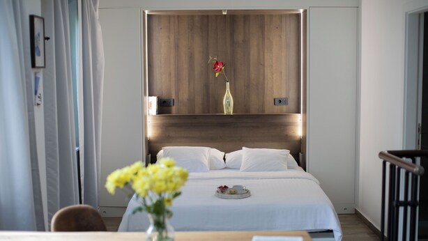 A hotel room with a bed, a bedside table with a tray of food, a vase with flowers, and a wooden headboard.