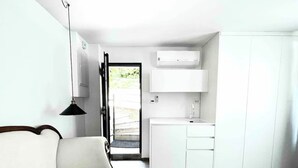 A modern living room with a sofa, a wooden coffee table, a pendant light, and a view of a kitchen with white cabinets and an air conditioning unit.
