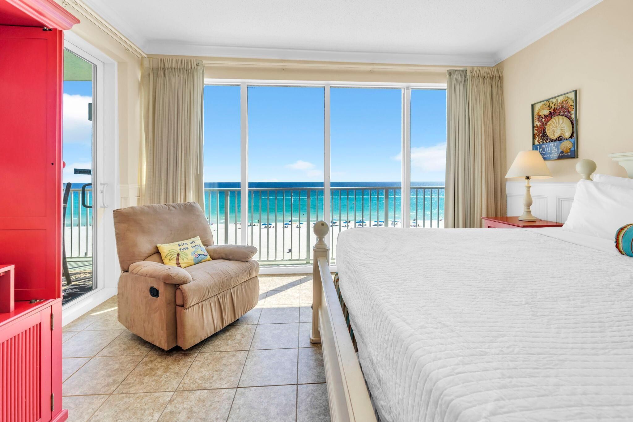 A hotel room with a bed, a reclining chair, a red cabinet, a nightstand, a lamp, and a large window with a view of the ocean.