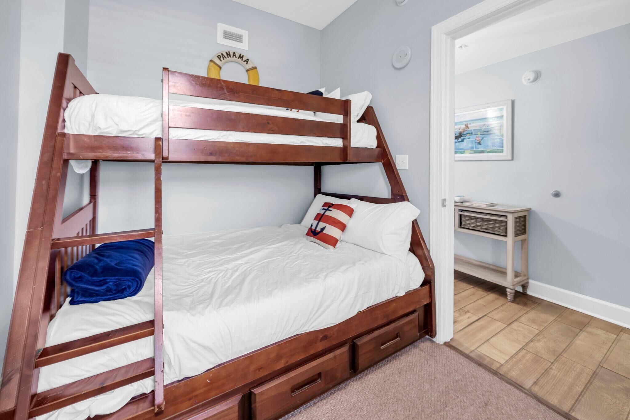 A bunk bed with white bedding, a blue blanket, a Panama sign, a small table with a basket, and a framed picture on the wall.