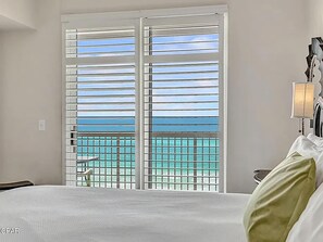 A hotel room with a bed, a nightstand, a lamp, and a view of the ocean through a balcony with metal bars. - Sunrise Beach #1010 (Panama City Beach)