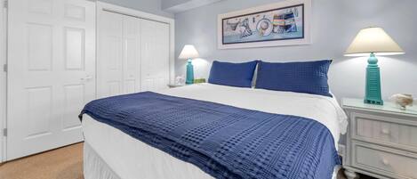 Un dormitorio con una cama grande, paredes blancas, un cuadro, dos lámparas de noche y un aparador blanco.