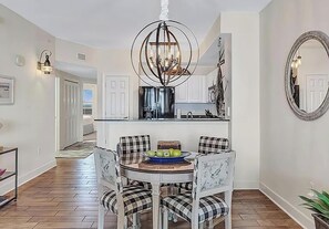 A dining area with a round table, plaid chairs, a chandelier, and a kitchen in the background. - Sunrise Beach #1010 (Panama City Beach)